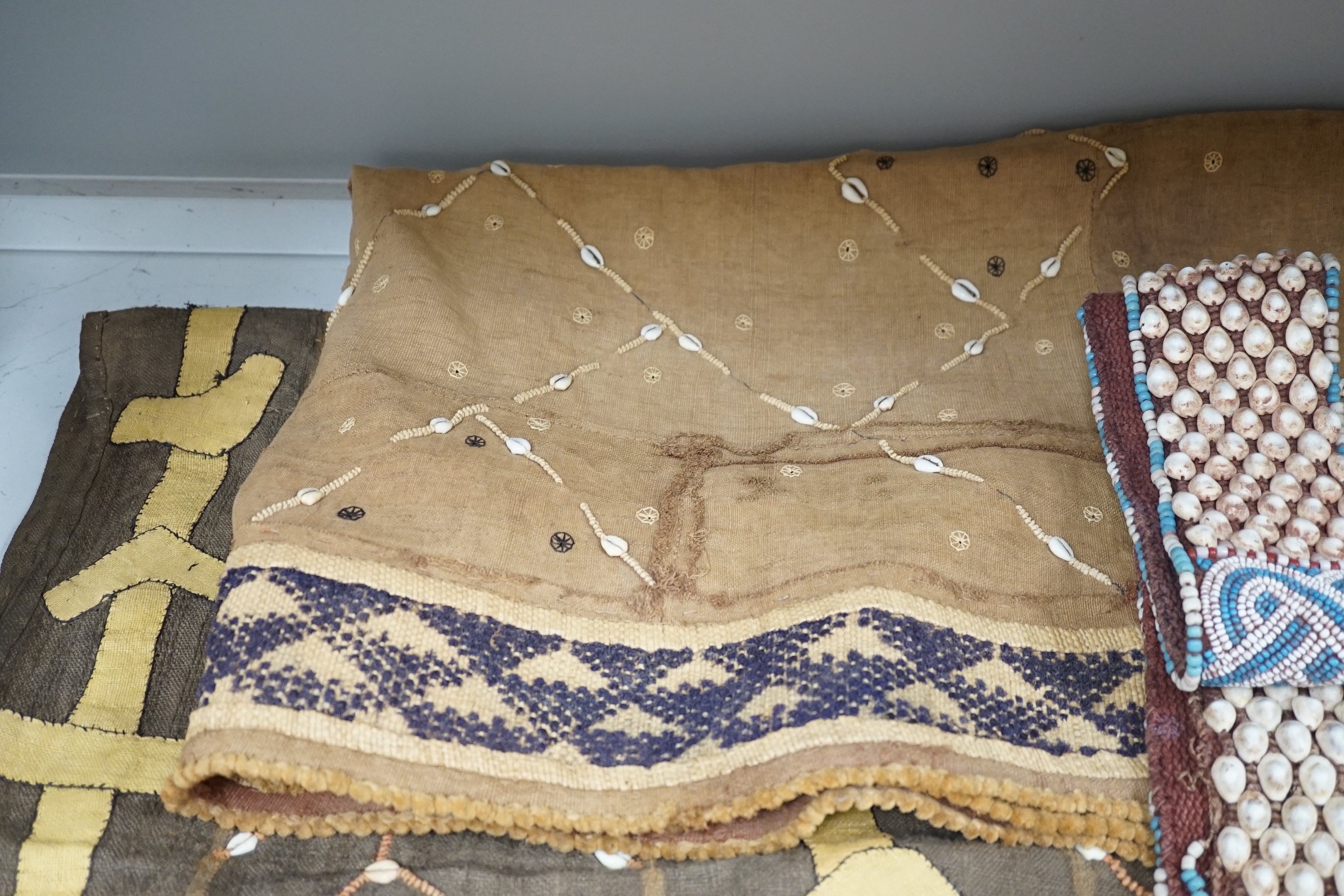 Three African tribal hangings, embroidered with coloured hand dyed threads and appliquéd shells on smaller handwoven cloth panels all sewn together to form the hangings, including a chieftains coloured bead worked and sh